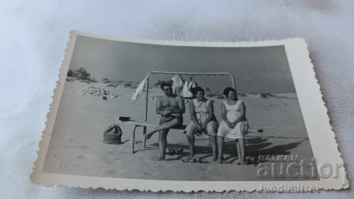 Fotografie cu un bărbat și două femei stând pe o bancă pe plajă