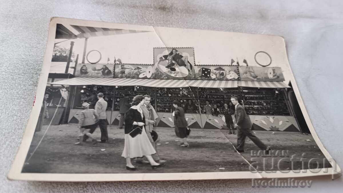 Fotografie Bărbați și femei în fața unui poligon de tragere într-un parc de distracții Fotografie Bărbați și femei în fața unui poligon de tragere într-un parc de distracții