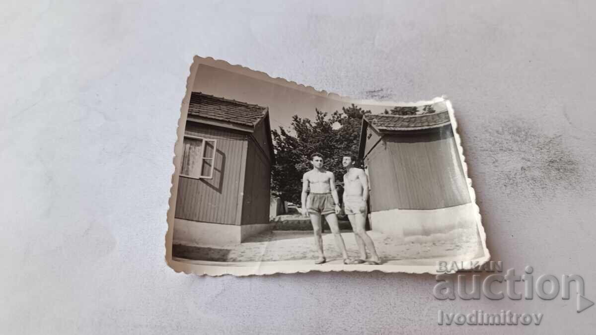 Fotografie doi bărbați în costume de baie în fața unor bungalouri