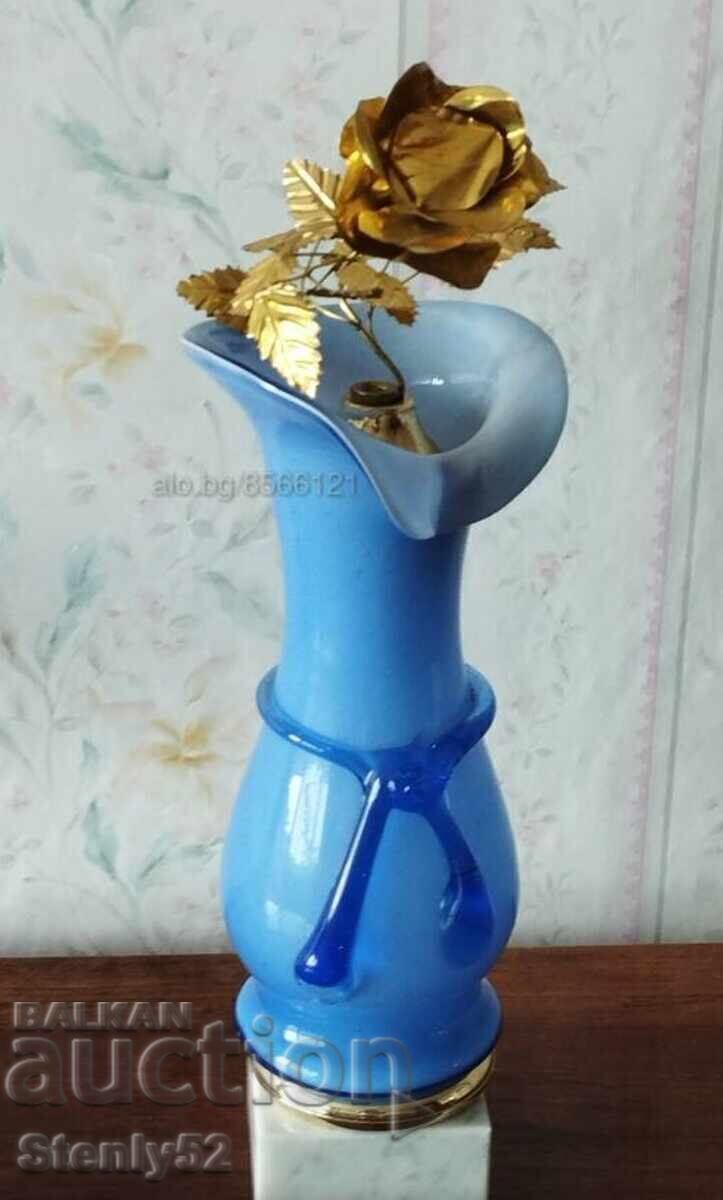 Vază din sticlă colorată cu un trandafir de metal pe un suport de marmură.