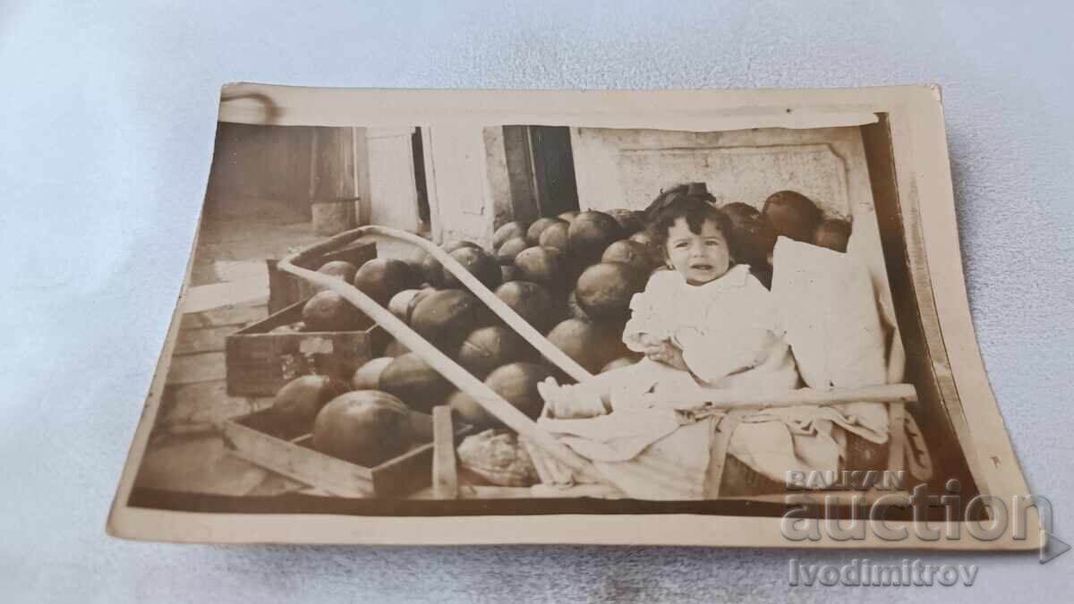 Photo of a little girl in a retro baby carriage in front of a pile of watermelons