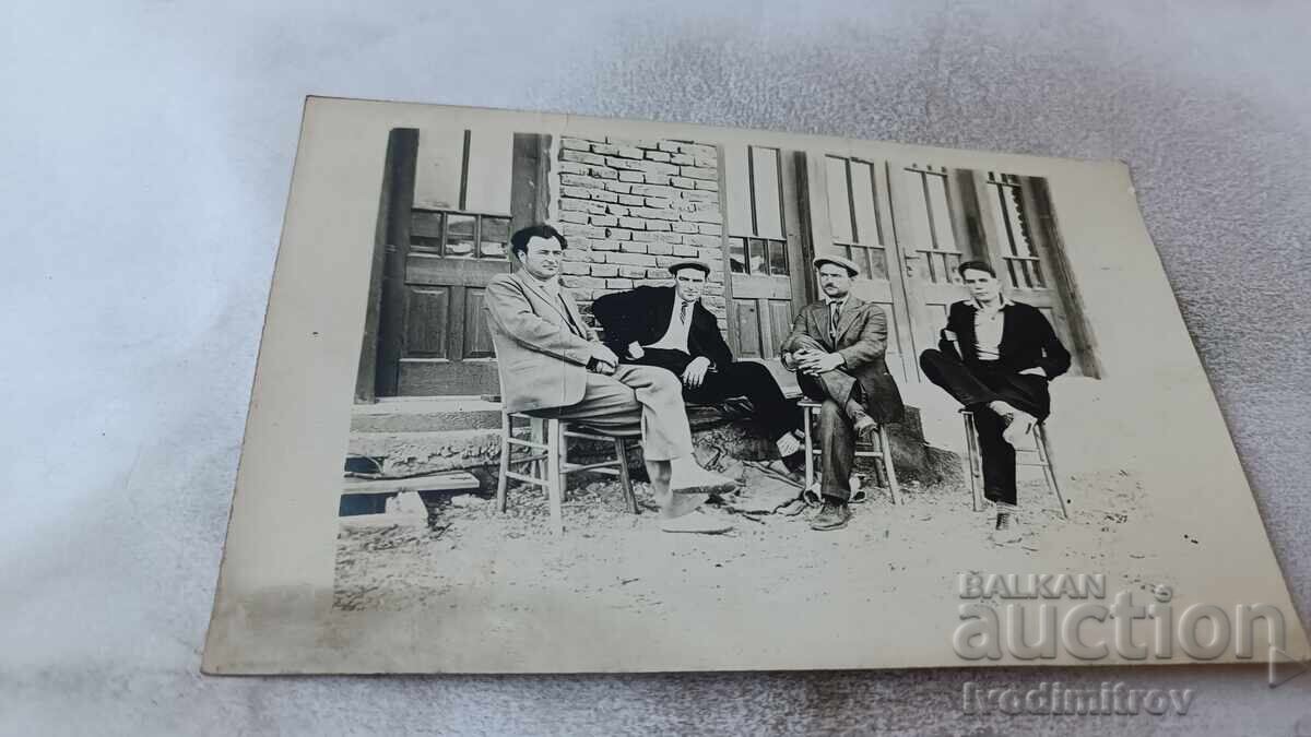 Fotografie cu patru bărbați stând pe scaune în fața unei clădiri Fotografie cu patru bărbați stând pe scaune în fața unei clădiri